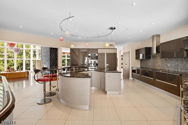 kitchen featuring light tile patterned floors, a center island with sink, tasteful backsplash, paneled built in fridge, and wall chimney range hood