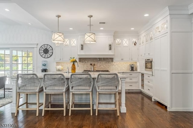 kitchen featuring backsplash, decorative light fixtures, white cabinets, oven, and an island with sink