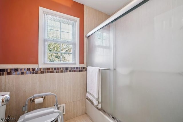 bathroom featuring tile walls, bath / shower combo with glass door, toilet, and a bidet