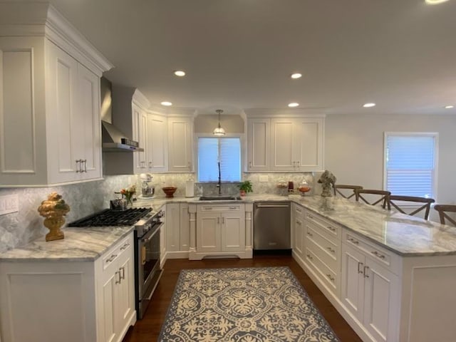 kitchen with wall chimney exhaust hood, white cabinetry, decorative light fixtures, kitchen peninsula, and stainless steel appliances