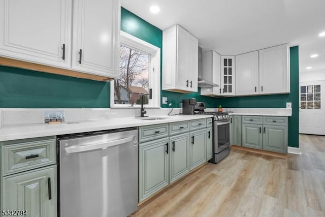 kitchen with white cabinets, appliances with stainless steel finishes, wall chimney range hood, sink, and light hardwood / wood-style flooring