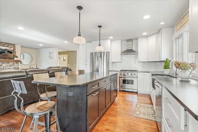 kitchen featuring decorative light fixtures, wall chimney range hood, a kitchen island, white cabinetry, and appliances with stainless steel finishes