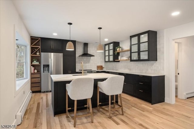 kitchen with decorative light fixtures, a kitchen island with sink, stainless steel fridge, baseboard heating, and wall chimney exhaust hood