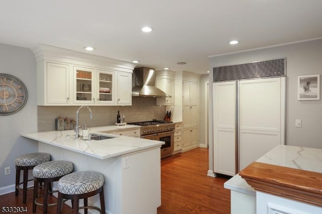 kitchen featuring wall chimney range hood, kitchen peninsula, sink, white cabinetry, and premium appliances