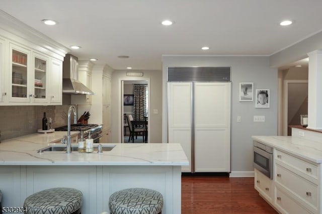 kitchen with white cabinetry, wall chimney range hood, built in appliances, backsplash, and light stone counters