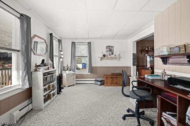 carpeted home office with a drop ceiling, wooden walls, and a baseboard radiator