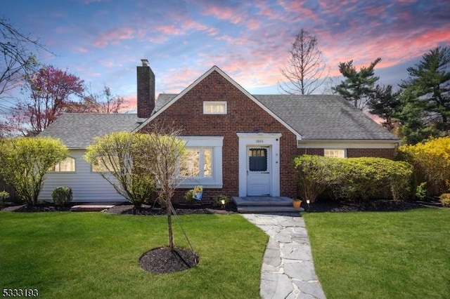 view of front facade featuring a shingled roof, a chimney, a yard, and brick siding