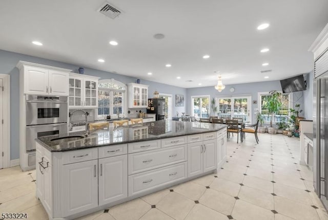 kitchen featuring white cabinetry, a center island with sink, dark stone counters, double oven, and sink