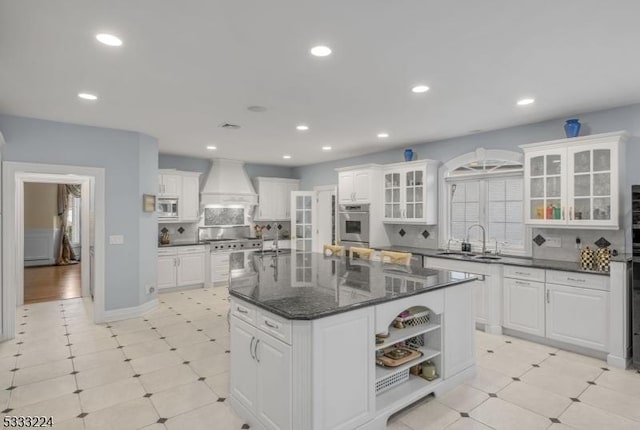 kitchen featuring tasteful backsplash, dark stone countertops, premium range hood, a kitchen island with sink, and white cabinets