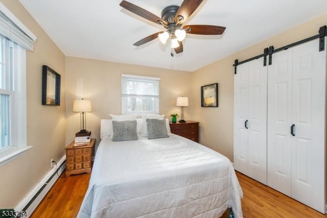 bedroom with ceiling fan, a baseboard heating unit, light hardwood / wood-style floors, a barn door, and a closet