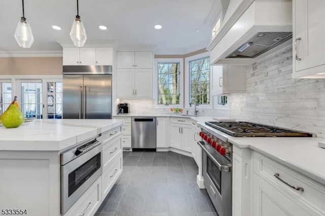 kitchen featuring built in appliances, custom exhaust hood, crown molding, white cabinets, and hanging light fixtures