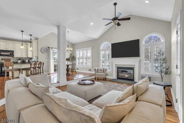 living room featuring light hardwood / wood-style flooring, ceiling fan, high vaulted ceiling, a tiled fireplace, and ornate columns