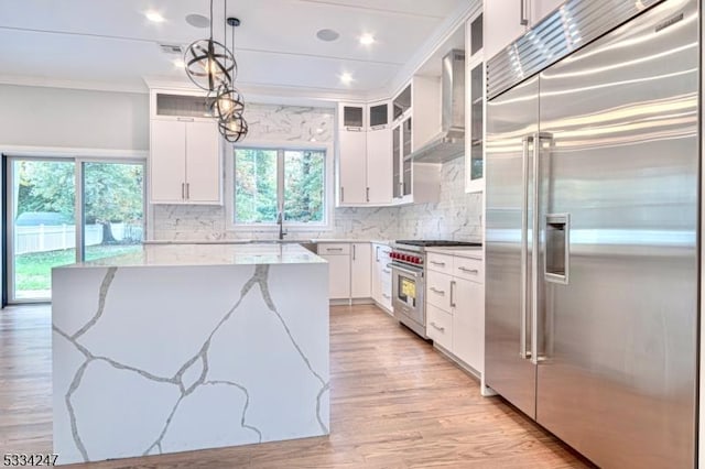 kitchen with decorative light fixtures, high quality appliances, wall chimney range hood, light stone countertops, and white cabinets