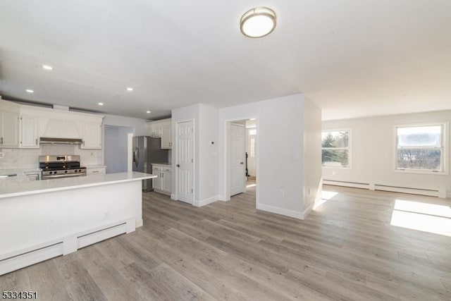 kitchen featuring wall chimney range hood, light hardwood / wood-style flooring, baseboard heating, appliances with stainless steel finishes, and white cabinets