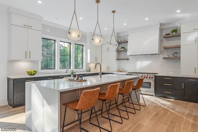 kitchen featuring wall chimney range hood, a kitchen island with sink, high end range, light hardwood / wood-style floors, and white cabinets