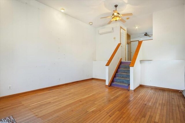 unfurnished living room featuring a wall unit AC, ceiling fan, light wood-type flooring, baseboards, and stairs