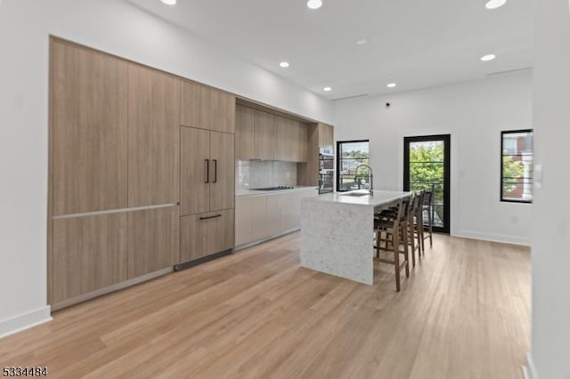 kitchen featuring a breakfast bar, an island with sink, black electric stovetop, light hardwood / wood-style floors, and decorative backsplash