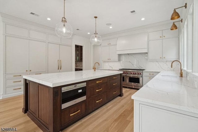 kitchen featuring an island with sink, premium range hood, sink, range with two ovens, and white cabinets