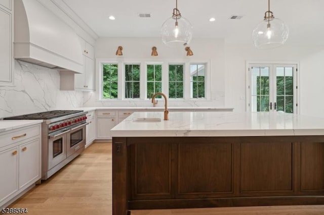 kitchen featuring range with two ovens, white cabinets, sink, custom exhaust hood, and a kitchen island with sink