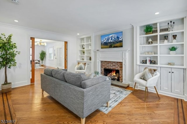 living room with a brick fireplace, built in shelves, ornamental molding, and light hardwood / wood-style floors