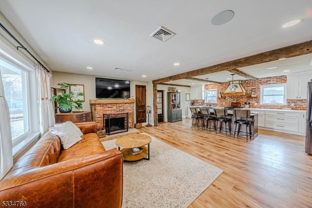 living room featuring beamed ceiling, a brick fireplace, a wealth of natural light, and light hardwood / wood-style floors