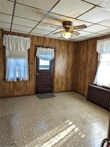 interior space featuring a drop ceiling, radiator, ceiling fan, and wood walls