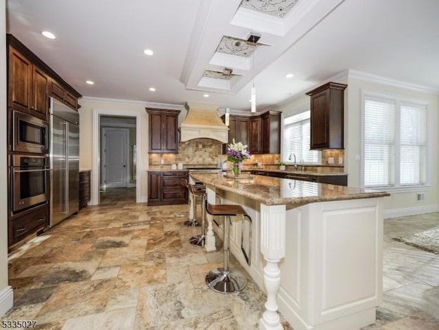 kitchen featuring a breakfast bar, built in appliances, a kitchen island, custom range hood, and light stone countertops