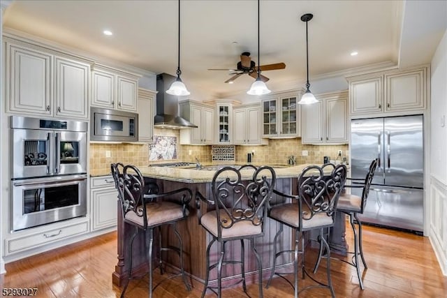 kitchen with wall chimney range hood, a kitchen island with sink, built in appliances, light stone counters, and decorative light fixtures