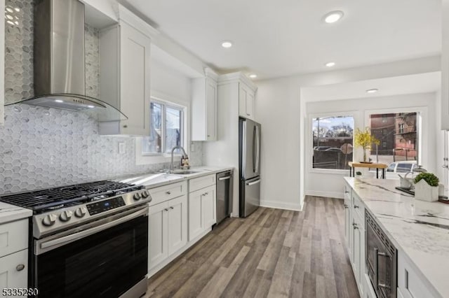 kitchen featuring appliances with stainless steel finishes, white cabinetry, sink, light stone countertops, and wall chimney range hood