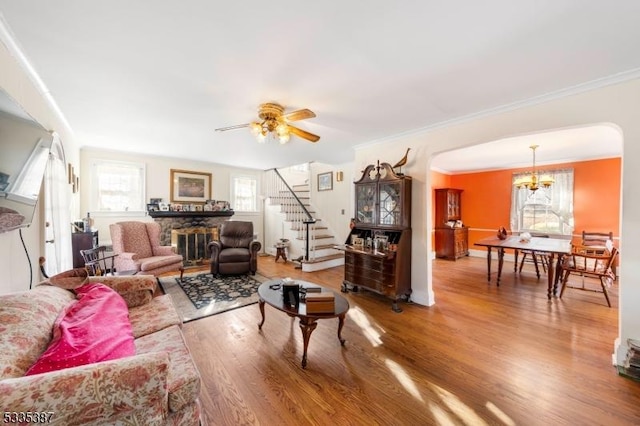 living room featuring ornamental molding, wood-type flooring, a stone fireplace, and ceiling fan with notable chandelier