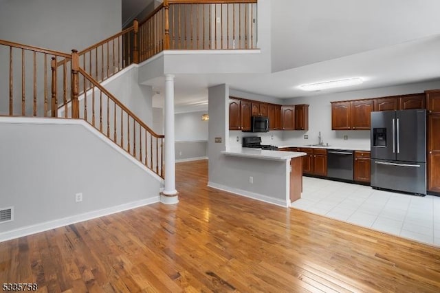 kitchen with a towering ceiling, stainless steel appliances, decorative columns, kitchen peninsula, and light wood-type flooring