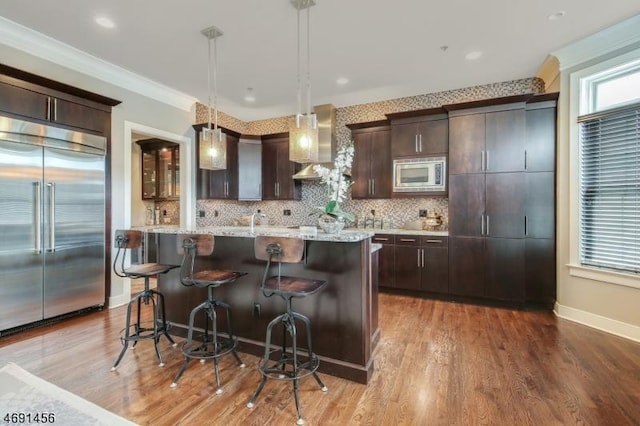 kitchen featuring wall chimney exhaust hood, built in appliances, a kitchen island with sink, and hanging light fixtures