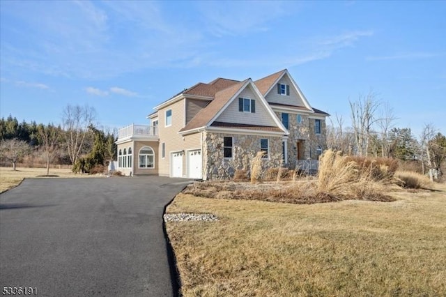 view of front of home featuring aphalt driveway, an attached garage, a balcony, stone siding, and a front lawn