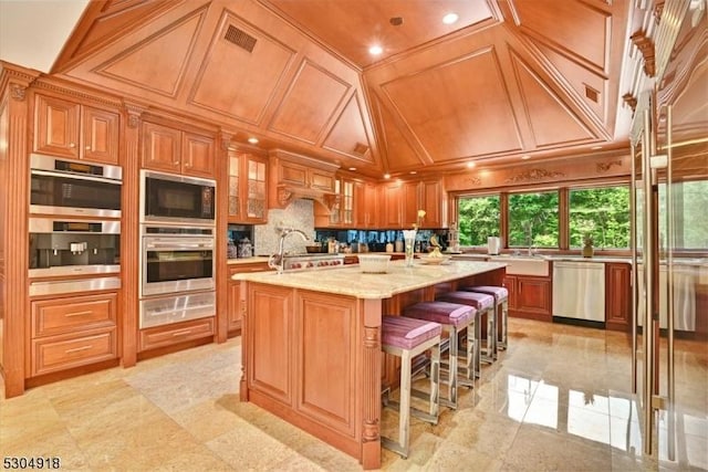 kitchen featuring appliances with stainless steel finishes, a kitchen breakfast bar, coffered ceiling, light stone countertops, and a center island with sink