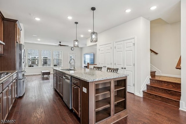 kitchen with dark brown cabinetry, appliances with stainless steel finishes, pendant lighting, light stone countertops, and a kitchen island with sink