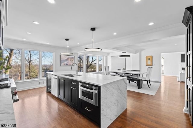 kitchen with sink, appliances with stainless steel finishes, hanging light fixtures, light stone counters, and a center island with sink