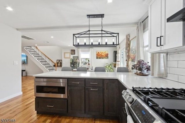 kitchen featuring stainless steel gas stove, dark brown cabinets, light wood-type flooring, hanging light fixtures, and wall chimney range hood