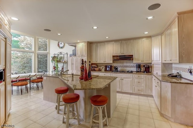 kitchen featuring light tile patterned flooring, a kitchen bar, tasteful backsplash, an island with sink, and light stone countertops