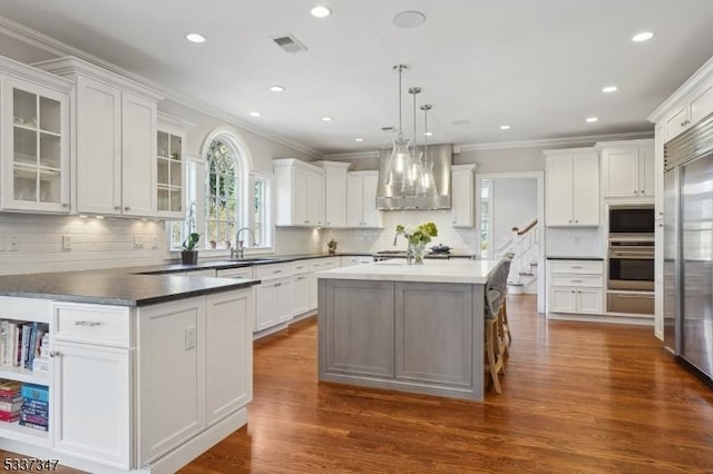 kitchen with dark countertops, a kitchen island, built in appliances, hanging light fixtures, and wall chimney range hood