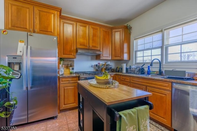 kitchen with brown cabinets, light tile patterned floors, appliances with stainless steel finishes, butcher block countertops, and under cabinet range hood