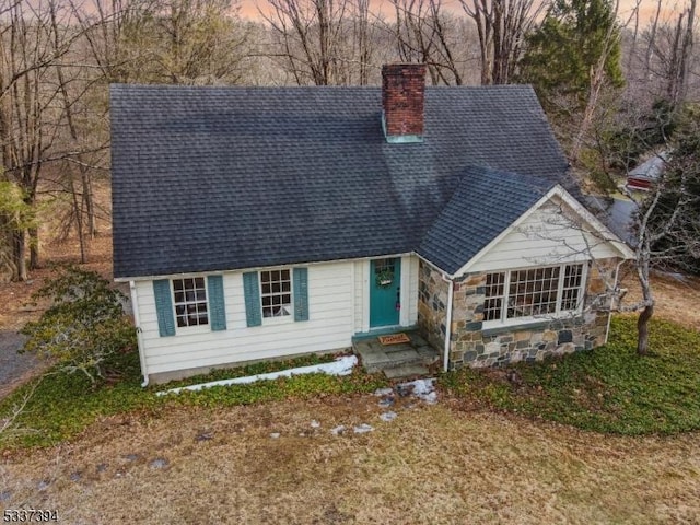 cape cod-style house with entry steps, stone siding, a chimney, and roof with shingles