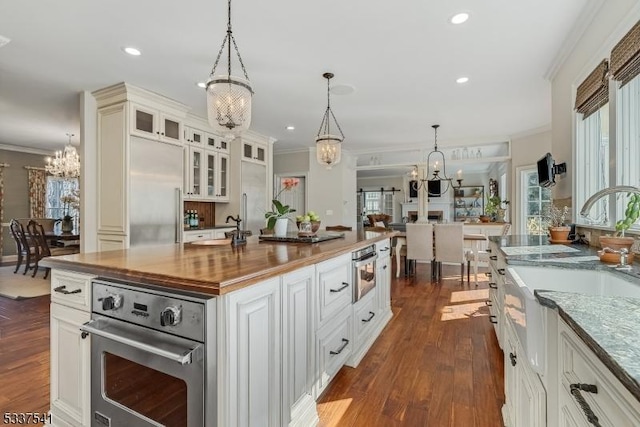 kitchen with a kitchen island with sink, a notable chandelier, stainless steel appliances, butcher block counters, and glass insert cabinets