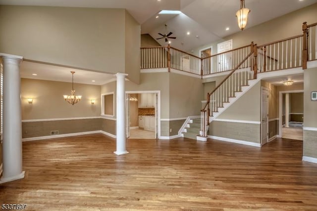 unfurnished living room featuring decorative columns, stairway, a towering ceiling, wood finished floors, and ceiling fan with notable chandelier