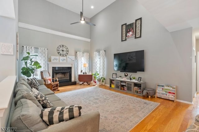 living room with a high ceiling, a premium fireplace, a ceiling fan, baseboards, and light wood-type flooring