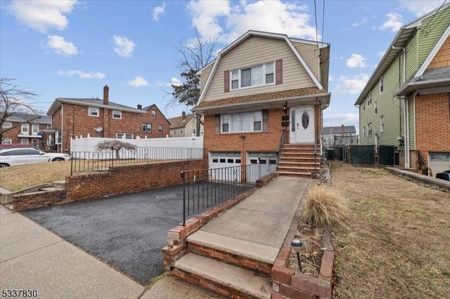 view of front of home featuring a gambrel roof, driveway, fence, an attached garage, and brick siding