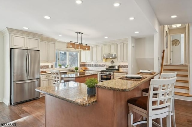 kitchen featuring light stone counters, stainless steel appliances, cream cabinets, a kitchen island, and under cabinet range hood