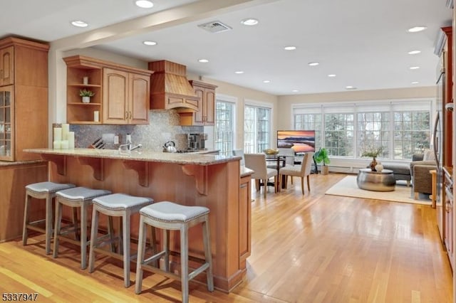 kitchen with a breakfast bar area, light wood finished floors, custom exhaust hood, visible vents, and light stone countertops