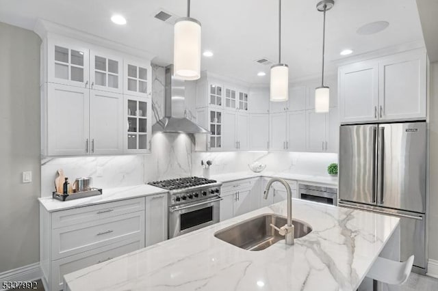 kitchen featuring premium appliances, decorative light fixtures, white cabinetry, a sink, and wall chimney range hood