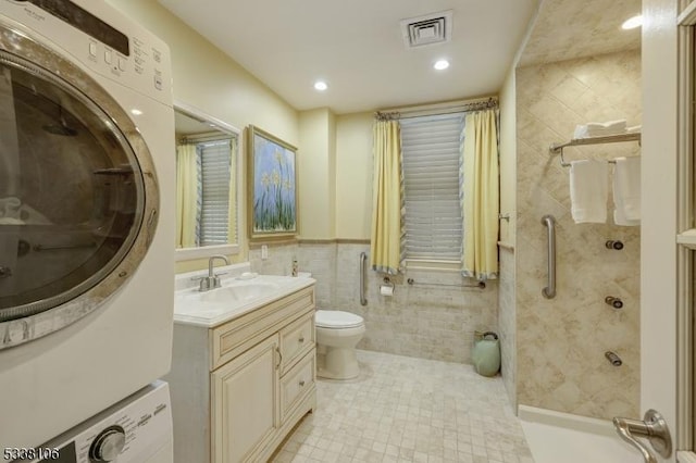 bathroom featuring toilet, tile walls, a tile shower, vanity, and stacked washing maching and dryer