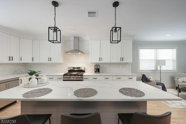 kitchen featuring wall chimney exhaust hood, light stone counters, hanging light fixtures, stainless steel range with gas cooktop, and white cabinets